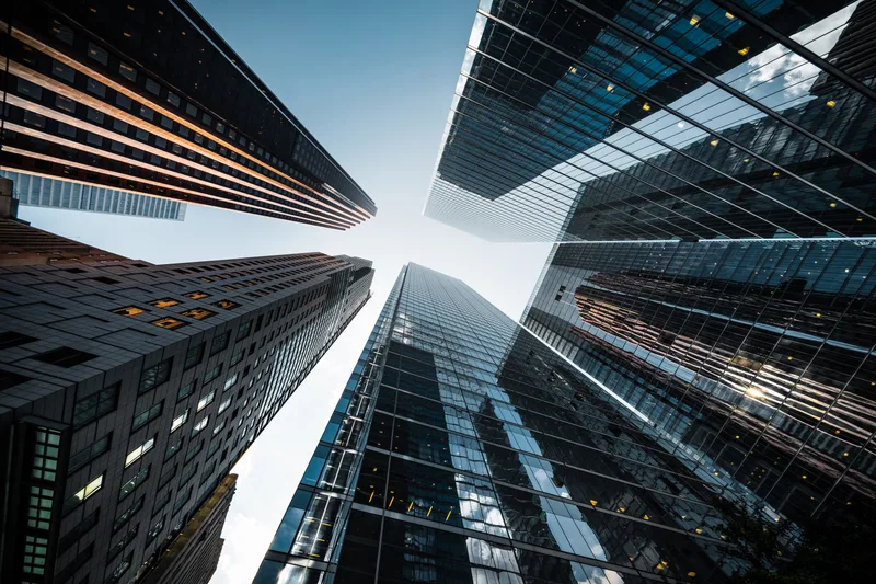 Looking straight up between glass office towers toward a pale blue sky