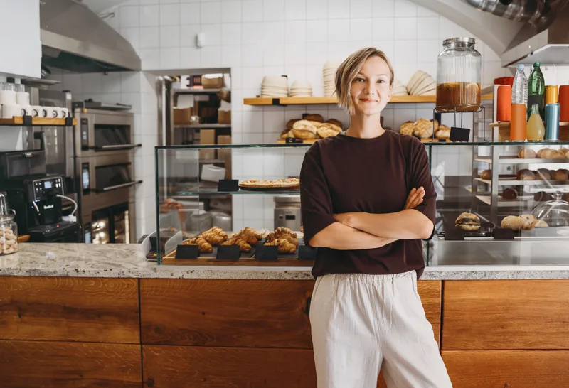 A bakery owner stands with arms crossed in front of her pastry counter