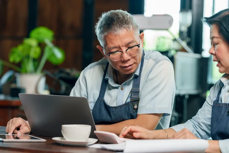 Two cafe owners in aprons go over figures on a laptop together at a table