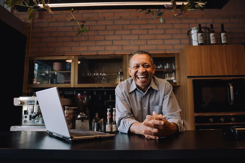 A smiling senior coffee shop owner leans on the counter of his cafe beside a laptop