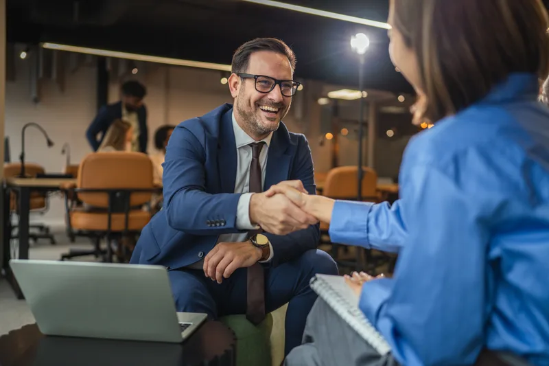 A lawyer in a blue suit greets a client with a handshake at a cafe table