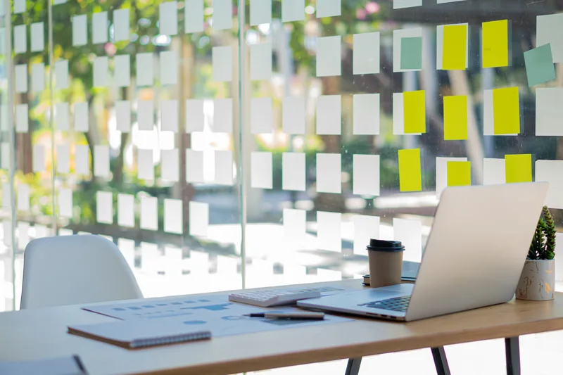 A desk with a laptop, notebook and coffee in front of a glass wall of sticky notes