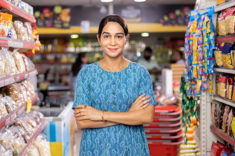 A grocery store owner stands with arms crossed in an aisle of her shop