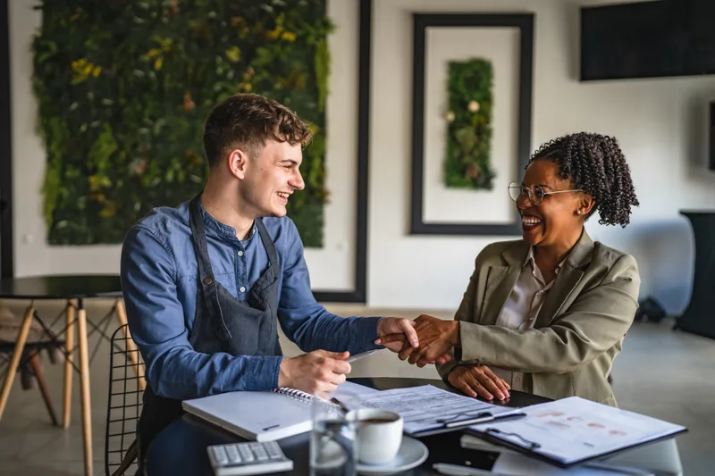 A cafe owner in an apron shakes hands with his business lawyer across a table of documents