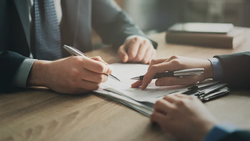 Two people at a table, one signing a document while the other points to the signature line