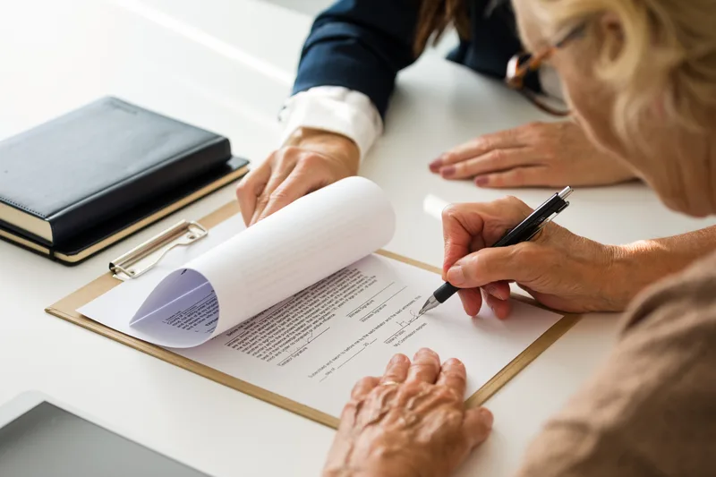 An older woman signing her will on a clipboard while her attorney holds the page steady
