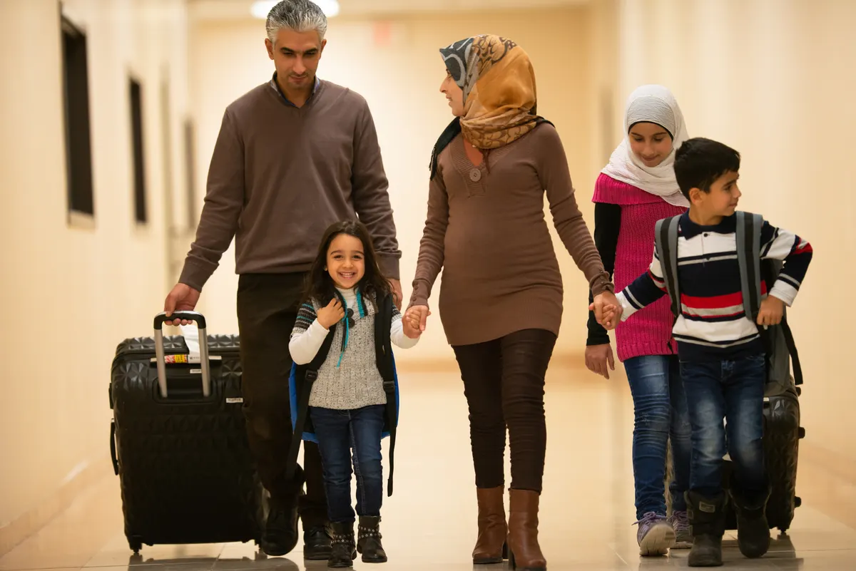 A family of five walks down a bright hallway with their suitcases, the youngest daughter smiling ahead of her parents