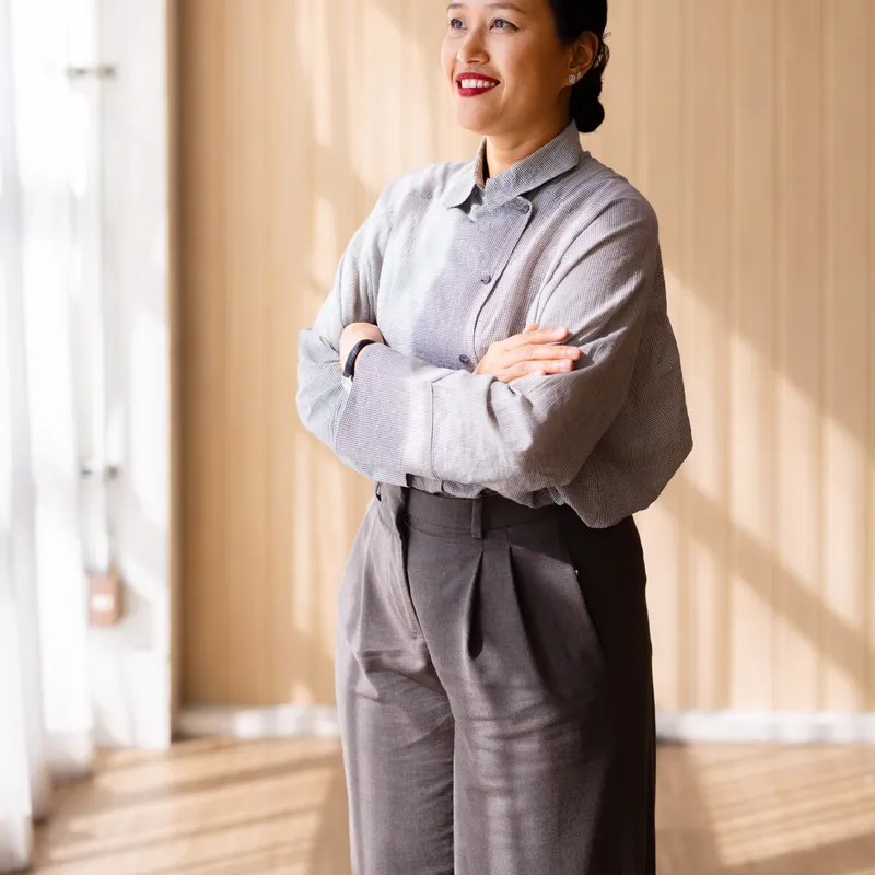 Mei-Ling Chow, an immigration consultant in a grey blouse, standing with her arms folded in a sunlit office