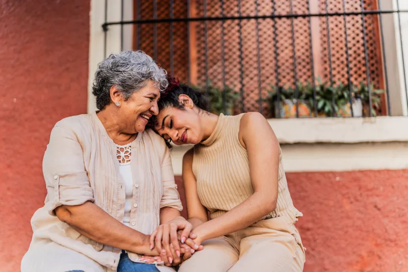 A grandmother and her adult granddaughter sit close together on front steps, holding hands and smiling