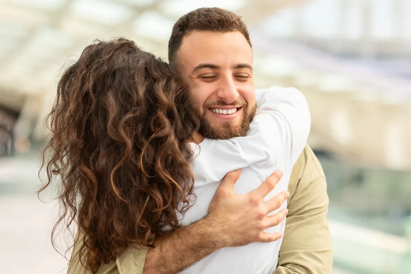 A smiling man hugs his wife tightly in a bright airport terminal after her flight lands