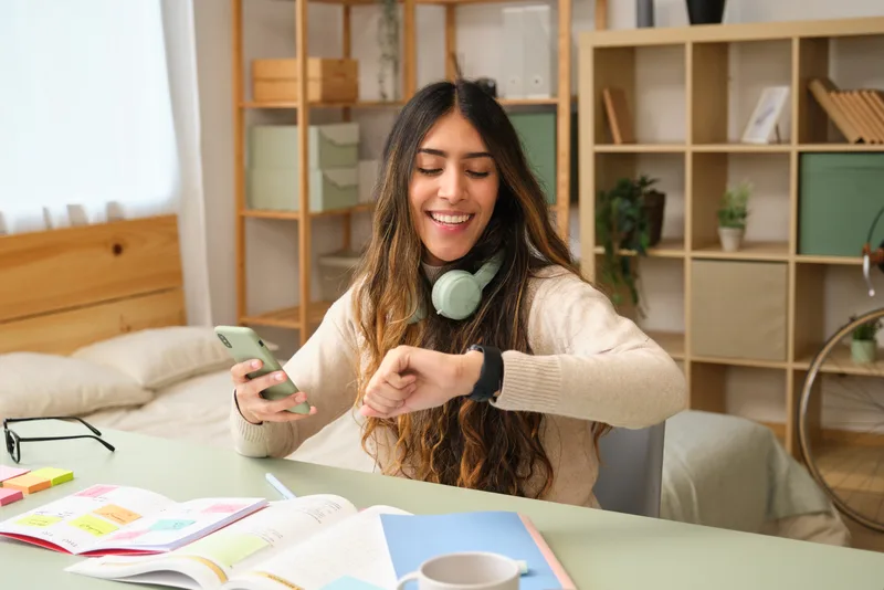 A young woman studying at home checks the time on her watch, notebooks open on the table