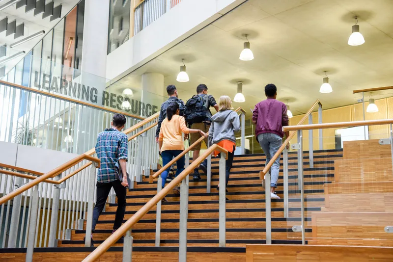 Students climb a wide wooden staircase inside a college library building