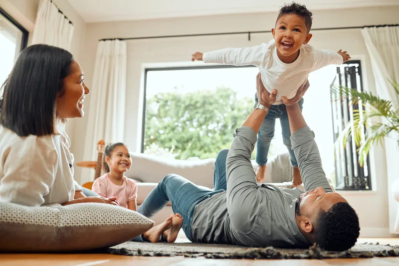 A parent lying on a rug lifting a laughing child into the air while family watches
