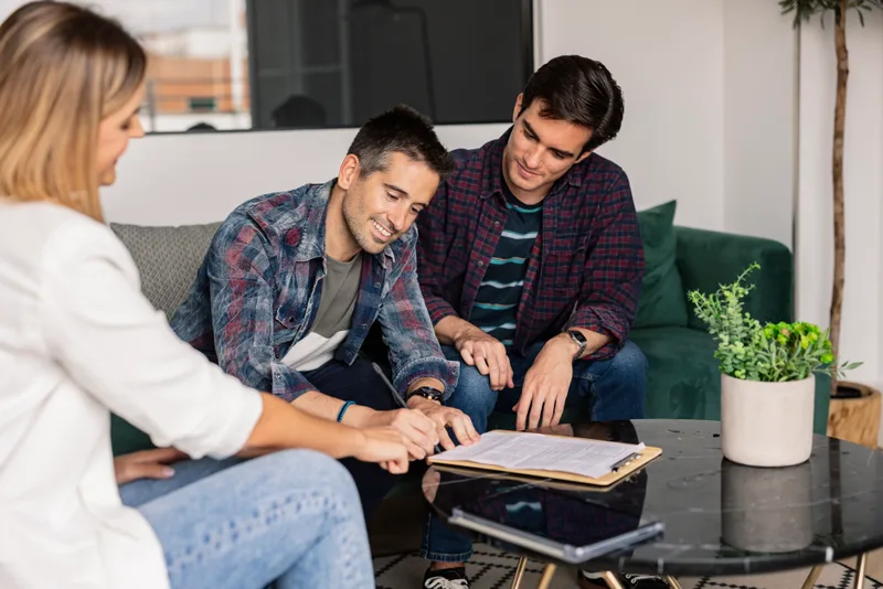 Two men signing papers at a coffee table while a lawyer explains the next page