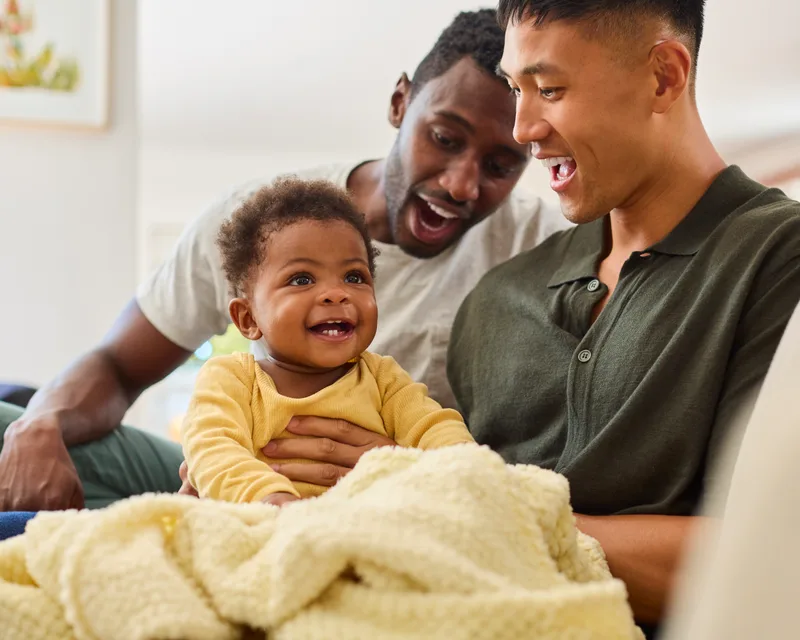 Two dads on a sofa smiling at their baby, who is laughing under a yellow blanket