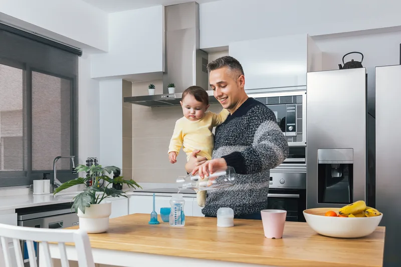 A father holding his baby in a bright kitchen while making a bottle