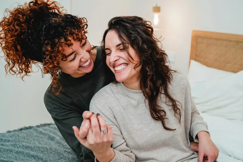 Two women laughing together at home, one showing a ring on her hand