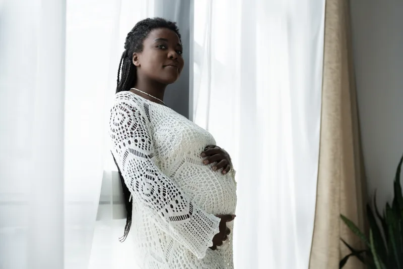 A pregnant woman in a white knit dress standing by a bright window