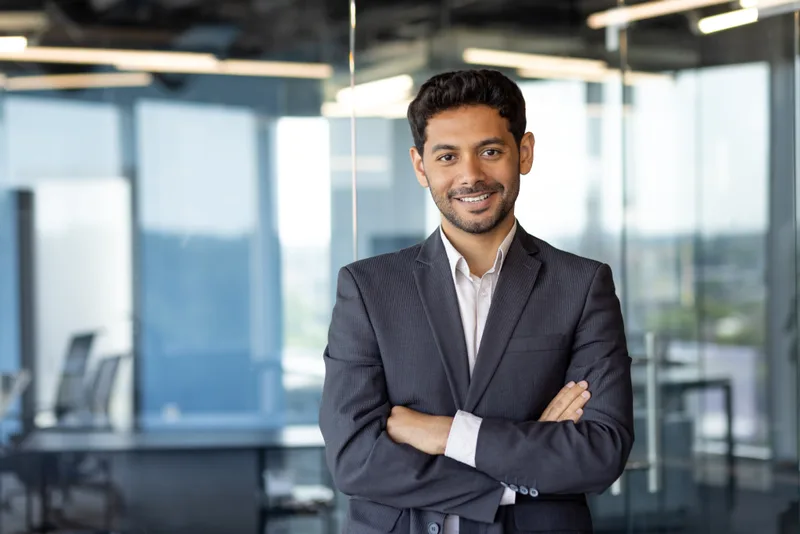 Arjun Mehta, a smiling attorney in a gray suit, standing with folded arms in a bright office