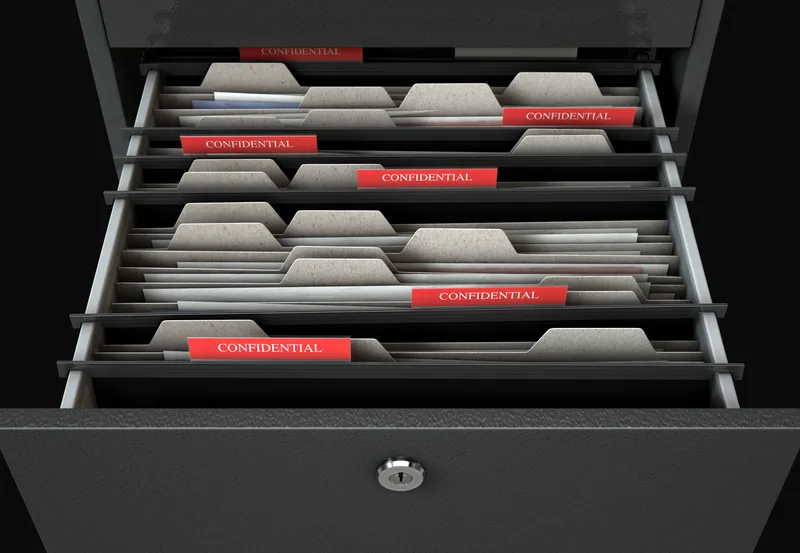 An open file drawer of folders with red tabs marked confidential