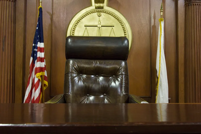 An empty judge's bench and leather chair in a wood-paneled courtroom with a flag beside it