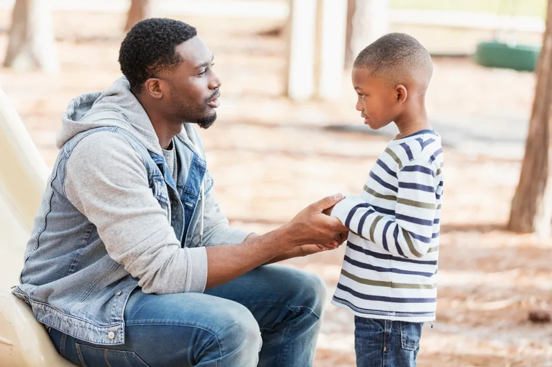A father sits at eye level with his young son at a playground, holding his hands while they talk