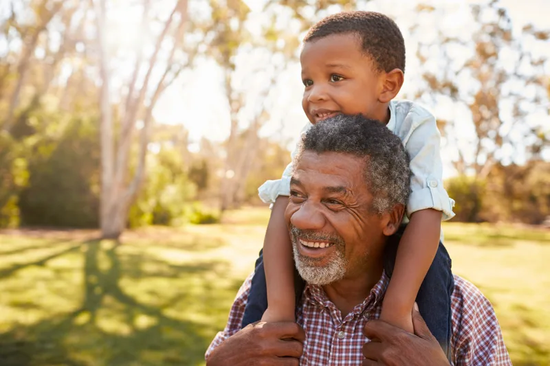 A laughing grandfather carries his grandson on his shoulders in a park