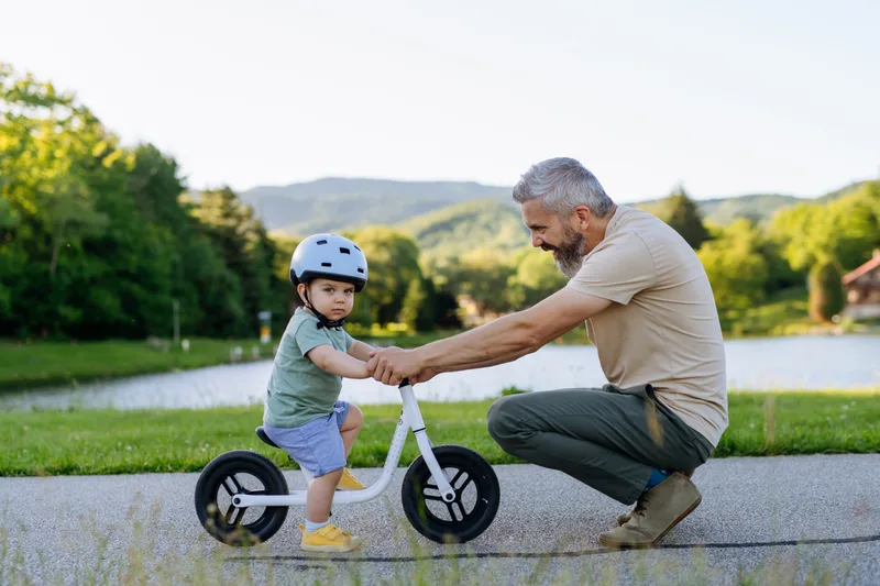 A father crouches beside his small son, steadying a balance bike on a path by a lake