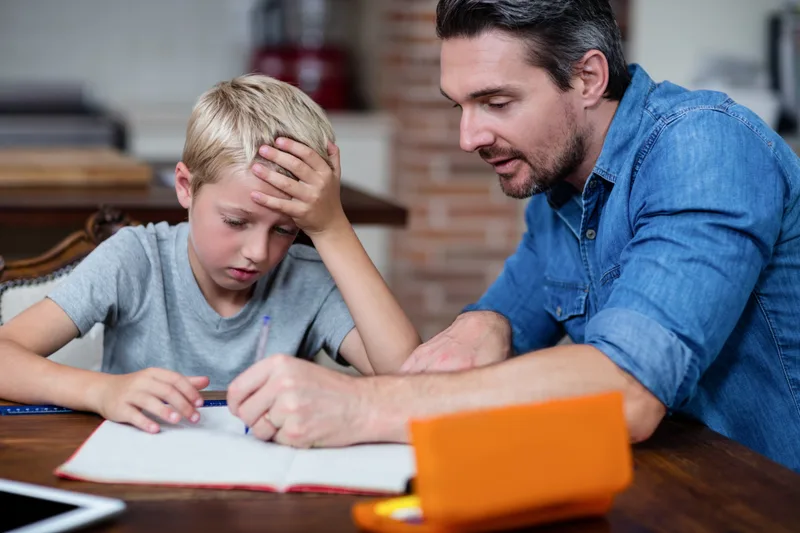 A father leans in to help his son with homework at the kitchen table