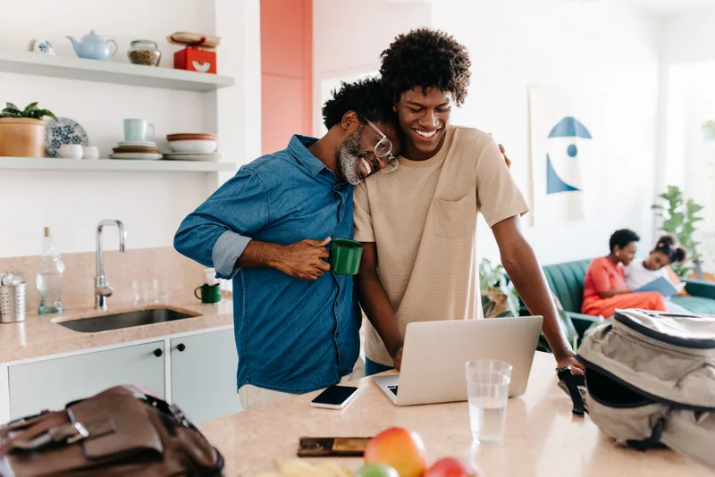 A father and his teenage son share a hug over a laptop in a bright kitchen before school