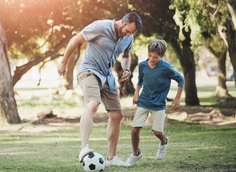 A father and son kick a ball across a park lawn in late afternoon light
