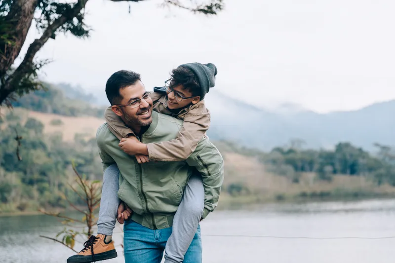 A smiling father carries his son on his back on a misty morning by a lake