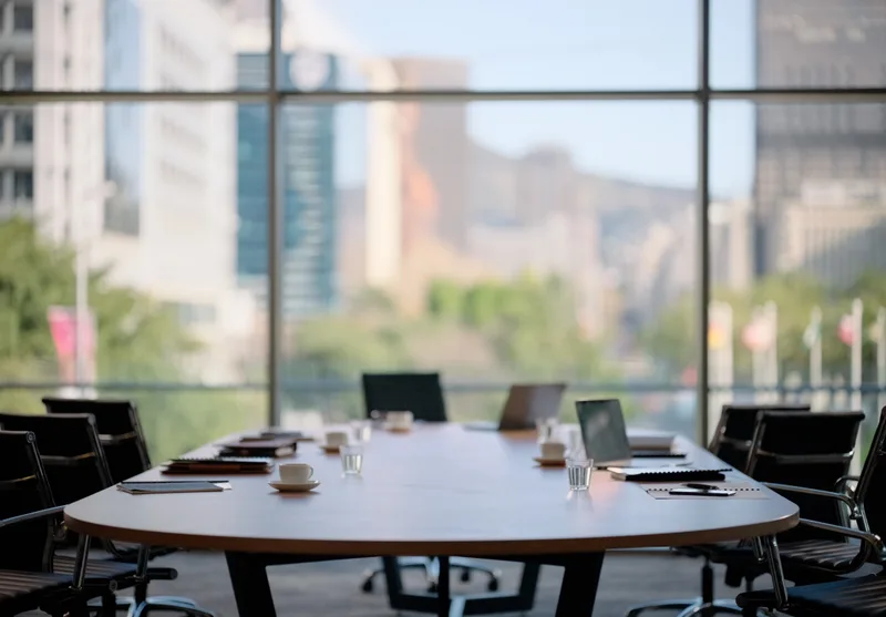 An empty conference table set with notebooks and laptops in front of tall windows over the city