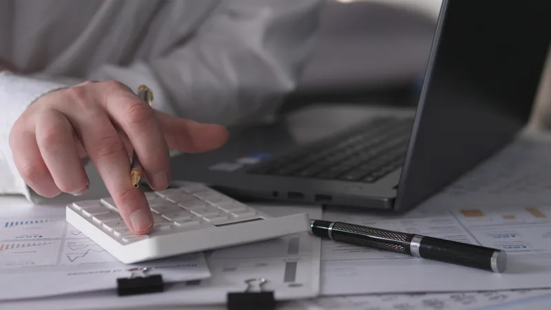 A hand works a desk calculator beside a laptop and printed financial statements