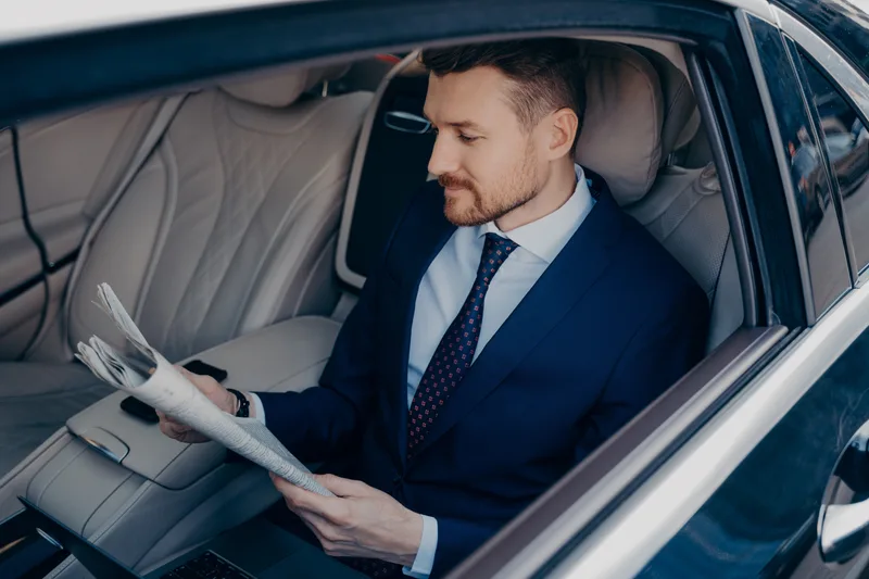 A man in a suit reads a newspaper in the back seat of a car