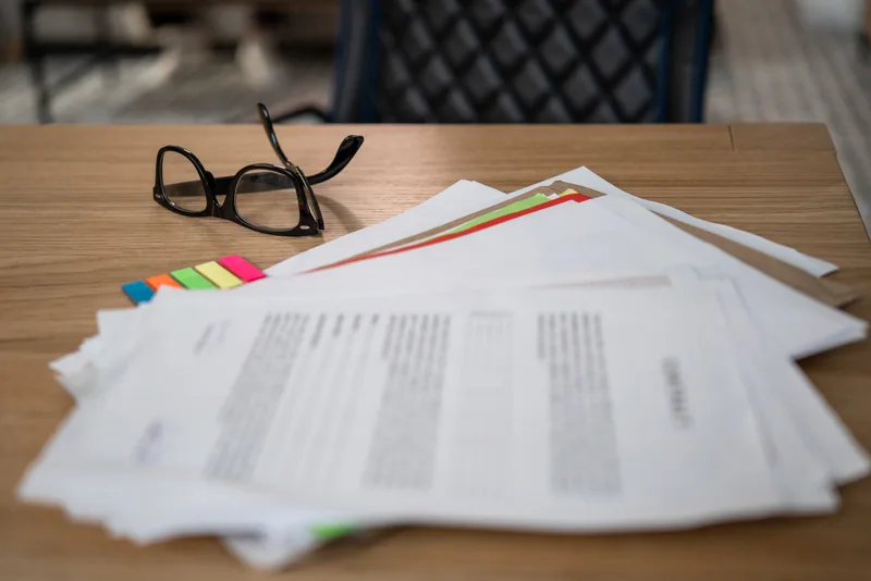 Printed records with colored index tabs spread across a wooden desk beside a pair of reading glasses