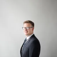 Tom Lindqvist, a smiling young man with glasses in a charcoal suit, photographed against a soft grey background