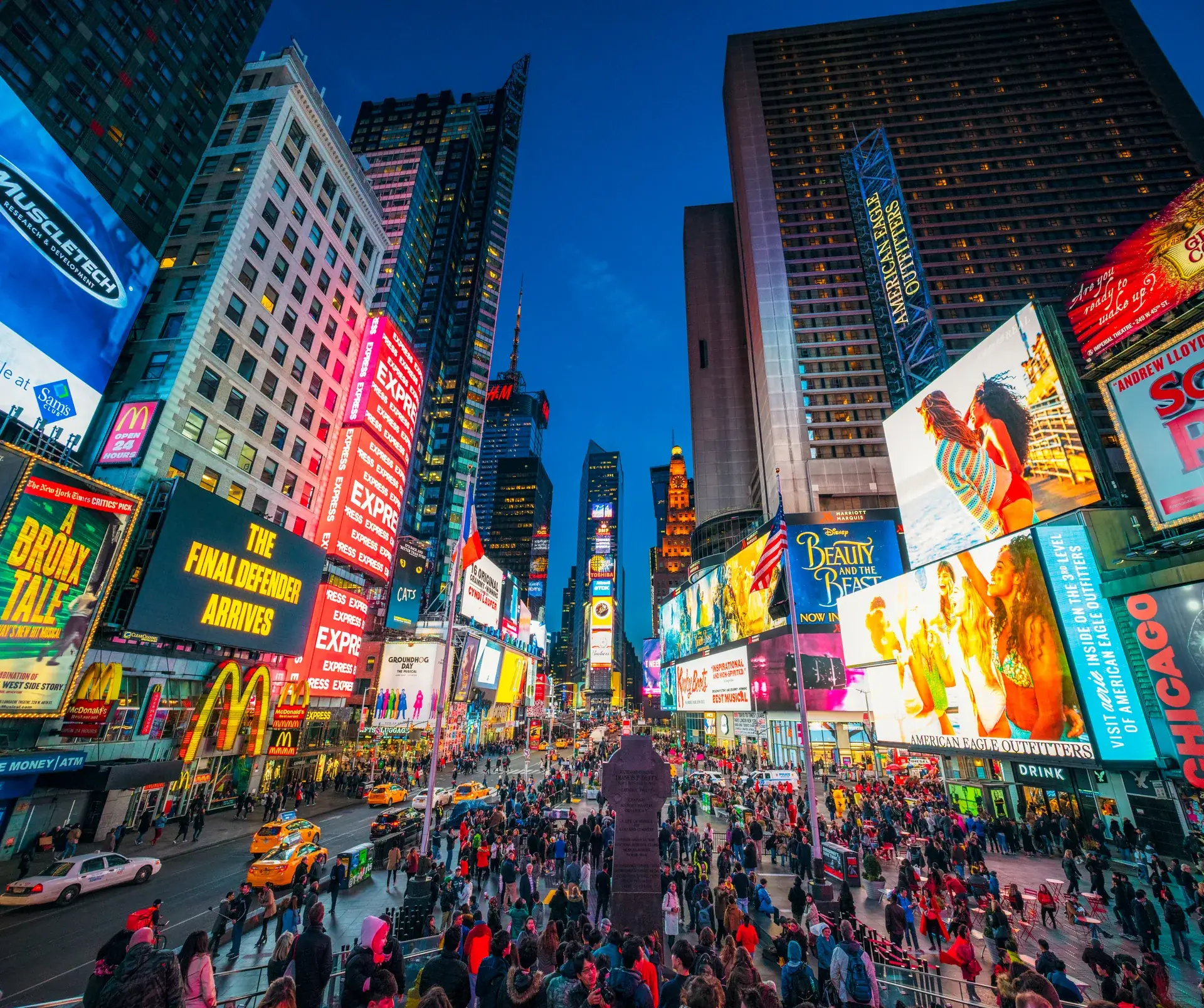 Multitudes cruzando Times Square al anochecer bajo las pantallas iluminadas — las calles de Nueva York que nuestros clientes recorren cada día
