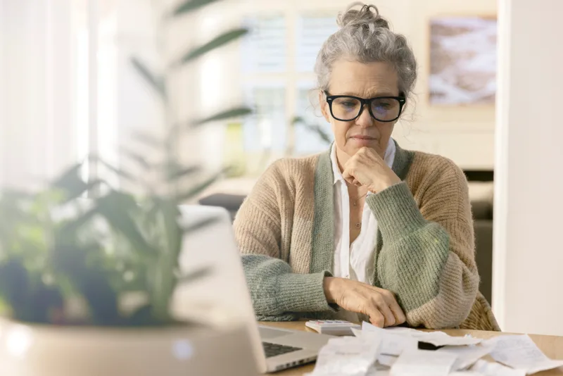 A woman with grey hair and glasses studies a pile of bills and statements at her table