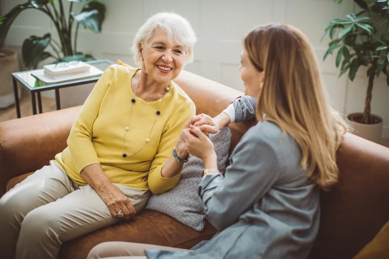 An adult daughter holds her smiling mother's hands as they talk together on a living room sofa