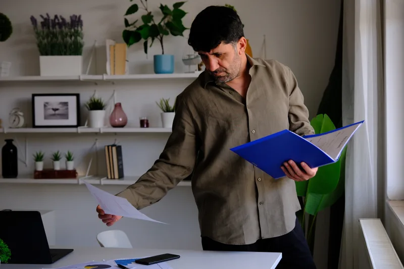 A man standing at a desk at home sorts through a blue folder of financial papers