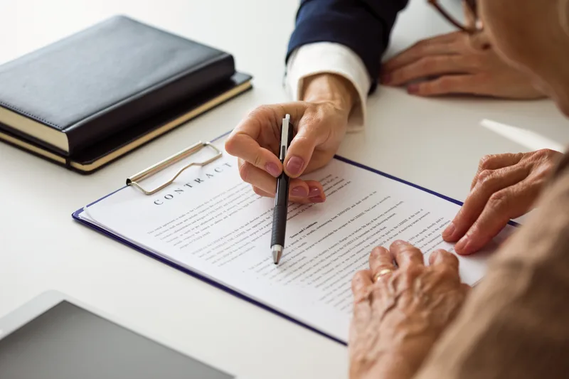 An older client signs a document on a clipboard while an attorney points to the signature line