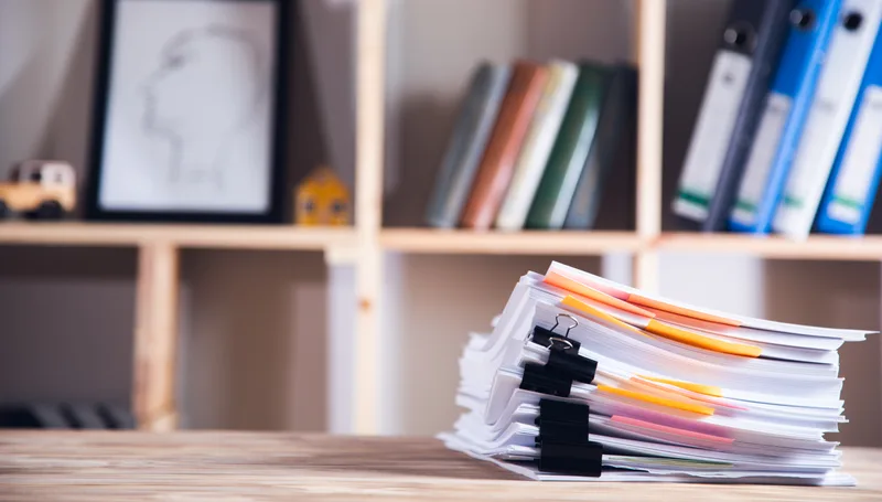 A tall stack of paper files held with binder clips on an office desk