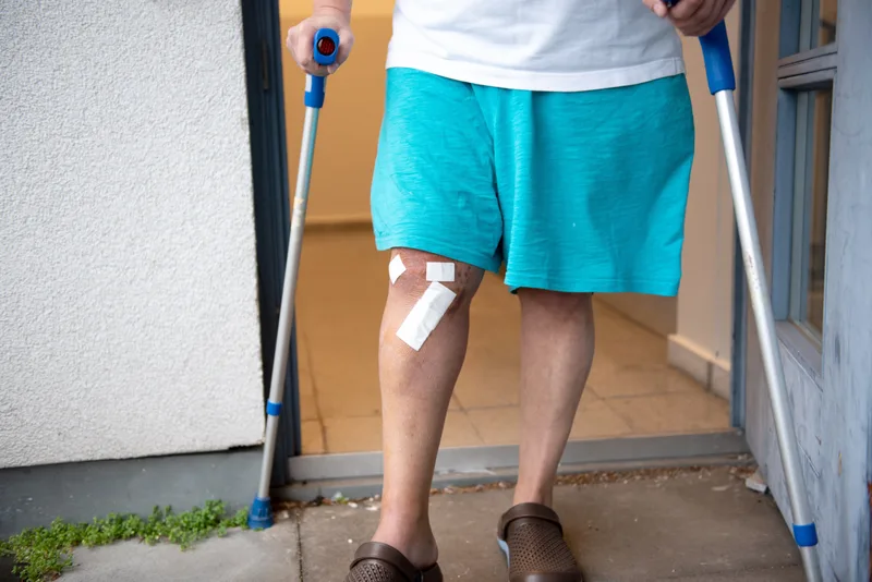 A man on crutches with a surgical dressing on his knee, standing in a doorway at home
