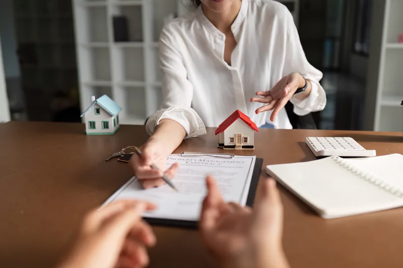 A client signs a document across a desk from an advisor, with two small model houses and a calculator on the desk