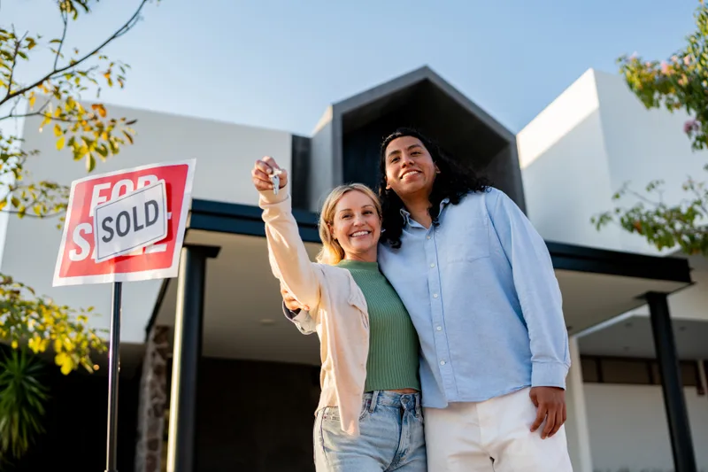 A smiling couple holds up a house key beside a sold sign in front of a modern home