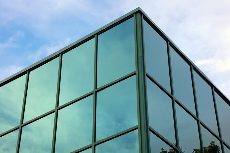 Looking up at the corner of a glass office tower, sky and clouds reflected in its green-tinted curtain wall