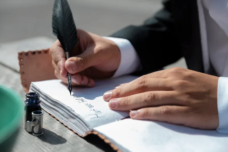 Close-up of hands in a dark suit writing in a bound notebook with a quill pen, an inkwell standing beside the page