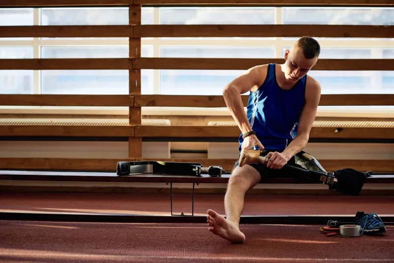 A young man sitting on a bench in a sports hall adjusts his prosthetic leg