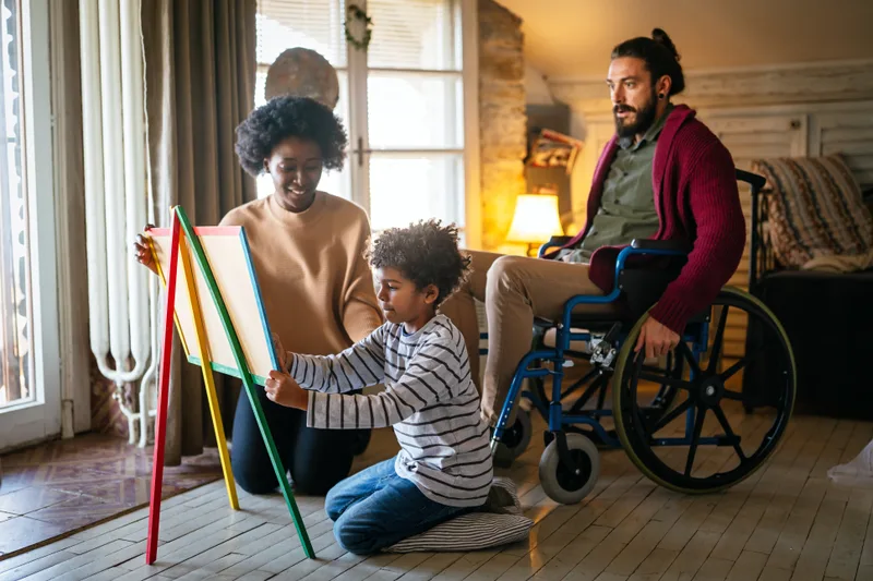 A father in a wheelchair watches his young son paint at an easel while the boy's mother kneels beside them in a warm living room